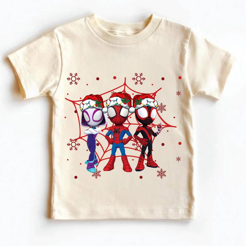 May include: Cream-colored t-shirt featuring three cartoon Spider-Man characters wearing Santa hats, set against a red spiderweb design with snowflakes. The characters are in their signature costumes. A festive holiday-themed graphic tee.