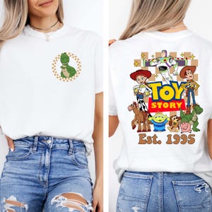 May include: White t-shirt featuring a green dinosaur graphic inside a brown and white checkered circle on the front. The back of the shirt displays a colorful Toy Story graphic with the text "Toy Story Est. 1995".