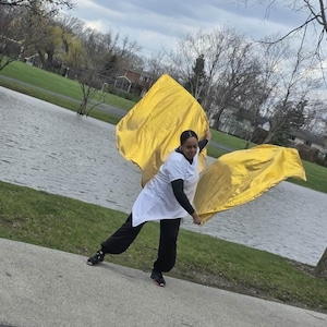 May include: A person is holding two large, flowing, golden yellow flags. They are wearing a white tunic over a black long-sleeved shirt and black pants. The background includes a body of water, green grass, and a cloudy sky.