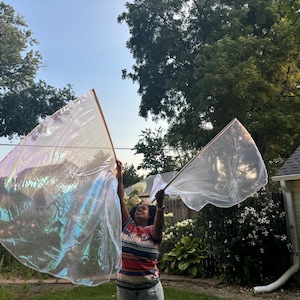 May include: A person holding two large, iridescent flags with wooden poles outdoors. The flags have a shimmering, translucent appearance, reflecting colors of blue, green, and purple. The person is wearing a striped shirt.