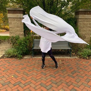 May include: A person is holding and moving large white fabric panels outdoors. The person is wearing a white top, patterned leggings, and black trainers. The background includes a brick walkway, a park bench, and trees.