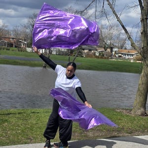 May include: A person is holding two large, shiny, purple flags outdoors. The flags are billowing in the wind. The person is wearing a white shirt, black pants, and black sleeves. The background includes a body of water and trees.