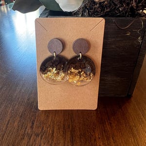 Puede incluir: Un par de pendientes hechos a mano. Cada pendiente presenta un pequeño perno redondo de madera oscura conectado a un colgante circular más grande y transparente. Los colgantes están rellenos de escamas de color marrón oscuro y dorado. Los pendientes se exhiben en una tarjeta marrón.