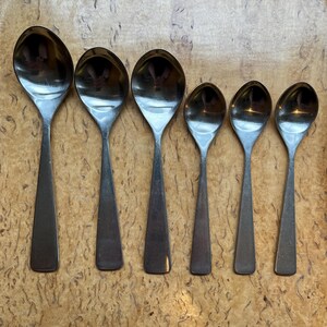 May include: Six stainless steel spoons of varying sizes are arranged on a wooden tray. The spoons have a simple, modern design with elongated handles and rounded bowls. The spoons are reflective, showing the surrounding environment.