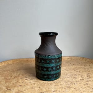 May include: A small, dark brown ceramic vase with a black rim and a cylindrical body. The body is decorated with horizontal bands of teal and black dotted patterns. The vase sits on a light brown wooden surface.