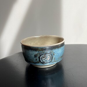 Peut inclure: Un petit bol en céramique fait à la main avec un glaçage bleu et beige. Le bol a un motif texturé sur le côté et un bord foncé. L'intérieur est de couleur beige clair et l'extérieur est bleu vif.