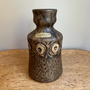 May include: A brown ceramic vase with a textured, mottled finish and a flared neck. The body features a repeating embossed circular pattern. A rectangular sticker on the front reads Lovemode Keramik HANDARBEJDE in black text on a gold background.