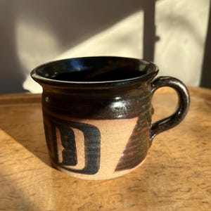 May include: A handcrafted ceramic mug with a dark brown glaze and a tan base. The mug features a black and tan geometric design. The handle is rounded and the rim is dark brown. The mug is on a wooden surface.