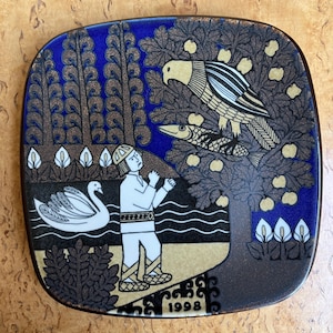 May include: A square ceramic decorative plate featuring a blue and brown illustration of a person standing near a swan in water, a tree with a bird holding a fish, and the year 1998 printed at the bottom. The design includes stylised plants.