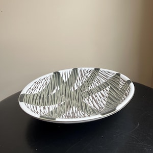 May include: Oval ceramic bowl with a white base and a black and grey linear pattern. The bowl has a black rim and is set against a neutral background. The design is a modern, abstract style.