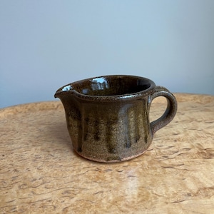 Peut inclure: Un petit pot à crème en céramique fait à la main avec un glaçage brun foncé. Le pot à crème a un bec verseur et une petite anse. Le glaçage a un effet dégoulinant, ajoutant de la texture à la pièce. Le pot à crème est posé sur une surface en bois.