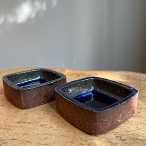 Peut inclure: Deux petits bols carrés en céramique, avec un extérieur marron foncé et un intérieur bleu profond. Les bols sont posés sur une surface en bois. Aspect rustique et fait main.