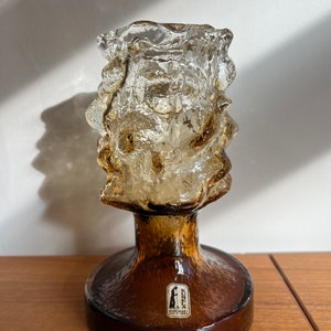 May include: A brown glass candle holder with a textured, organic design. The top portion has a rough, ice-like appearance, while the base is smooth and rounded. A small label is affixed to the base. The candle holder is on a wooden surface.