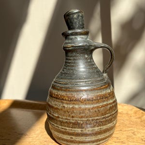Peut inclure: Une bouteille en céramique faite à la main avec un bouchon et une poignée gris foncé. La bouteille présente un motif rayé marron et beige. Cette bouteille est probablement pour l'huile ou le vinaigre et est un article de cuisine décoratif.