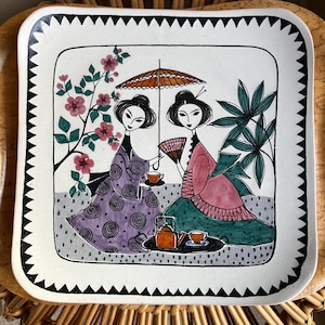 May include: A white ceramic serving tray with a black and white border. The tray features a hand-painted design of two women in traditional Japanese clothing, holding teacups and an umbrella, surrounded by flowers and plants.
