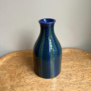 Peut inclure: Un vase en céramique bleu avec des rayures vertes verticales et de petits points noirs. Le vase a un col évasé et un corps arrondi. Le vase est posé sur une surface en bois brun clair.