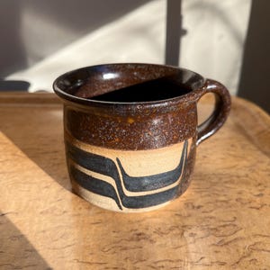 May include: A handmade ceramic mug with a dark brown speckled glaze. The mug features a tan band with two black curved lines. The handle is small and rounded. The mug is sitting on a wooden tray.