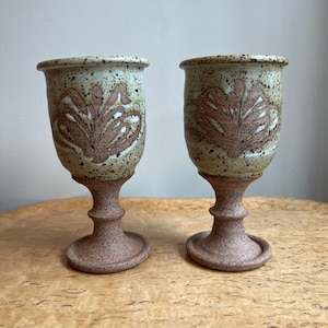 May include: Two ceramic goblets with a speckled, light green and brown glaze. Each goblet features a brown floral design and a pedestal base. The goblets are displayed on a wooden surface.