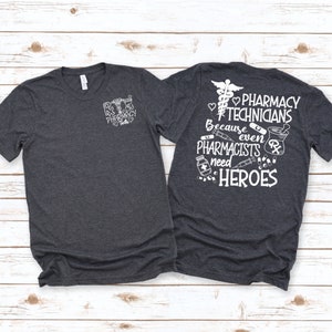Puede incluir: Una camiseta gris oscuro jaspeada con un gráfico blanco en la parte delantera que dice "Pharmacy Tech" y un gráfico blanco en la parte posterior que dice "Pharmacy Technicians Because even Pharmacists need HEROES".