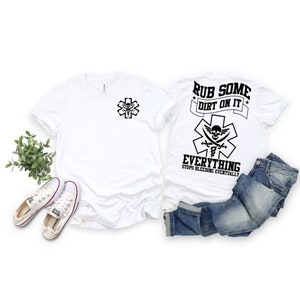 Puede incluir: Camiseta blanca con un símbolo médico negro y el texto "Rub some dirt on it. Everything stops bleeding eventually." en la parte posterior. La parte delantera de la camiseta tiene una versión más pequeña del símbolo médico.