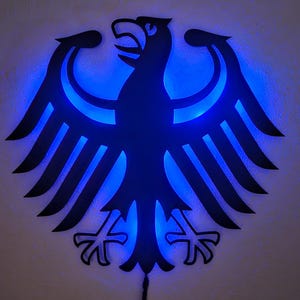 Könnte beinhalten: Schwarze Silhouette eines deutschen Adlers mit blauen LED-Lichtern, die durch die Ausschnitte scheinen. Der Adler ist ein Symbol Deutschlands.