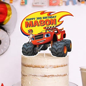 Puede incluir: Un adorno para pastel con un camión monstruo rojo, ojos azules y una cara sonriente. El adorno tiene un diseño de llama amarilla y naranja con el texto "HAPPY 3RD BIRTHAY MASON". Un personaje de dibujos animados está al lado del camión.