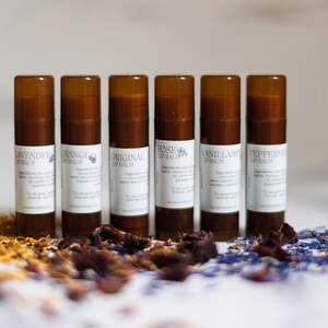 Puede incluir: Siete tubos marrones de bálsamo labial con etiquetas blancas. Las etiquetas tienen los nombres de los sabores: Lavanda, Naranja, Original, Rosa, Vainilla y Menta. Los tubos están dispuestos en una fila sobre una superficie blanca con pétalos de flores secas delante de ellos.