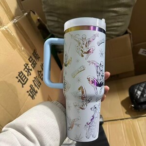 Peut inclure: Gobelet isotherme blanc avec plusieurs illustrations de Spider-Man. Le gobelet a une poign&eacute;e bleu clair et un couvercle blanc avec des accents dor&eacute;s et violets. Le mot "Spider-Man" est imprim&eacute; sur le gobelet.