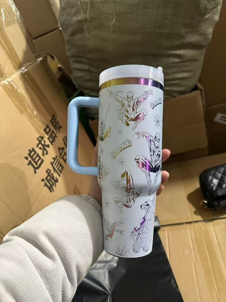Peut inclure: Gobelet isotherme blanc avec plusieurs illustrations de Spider-Man. Le gobelet a une poign&eacute;e bleu clair et un couvercle blanc avec des accents dor&eacute;s et violets. Le mot "Spider-Man" est imprim&eacute; sur le gobelet.
