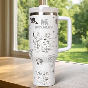 Puede incluir: Vaso t&eacute;rmico blanco con asa y tapa. El vaso presenta un dise&ntilde;o de un personaje alien&iacute;gena beb&eacute;, planetas, estrellas y flores. El logotipo de Stanley est&aacute; impreso en la parte delantera.
