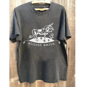 May include: A black short-sleeved t-shirt featuring a white graphic print of a bull standing on a hill with small flowers. Below the hill, the text "elliott smith" is printed in a white lowercase serif font. The shirt has a crew neckline.