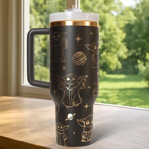 Puede incluir: Taza de viaje negra con asa, con ilustraciones doradas de un personaje de una popular serie de ciencia ficci&oacute;n. La taza tiene una tapa blanca con una banda dorada y un asa negra. El dise&ntilde;o incluye estrellas y planetas.