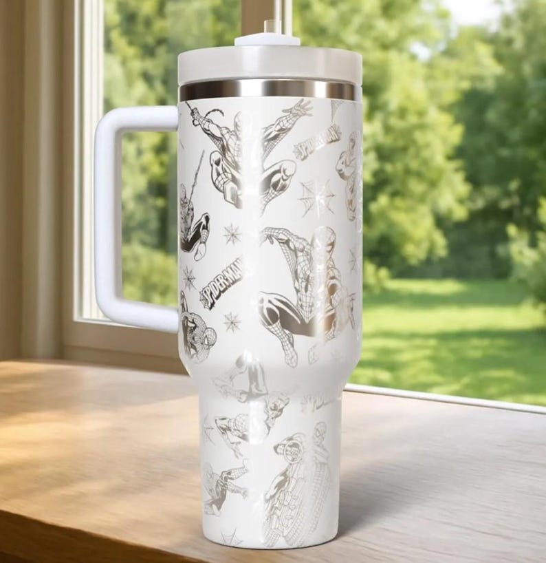 May include: White insulated tumbler with a handle, featuring a repeating pattern of Spider-Man illustrations in silver. The tumbler has a white lid and a silver band at the top. The word "SPIDER-MAN" is also visible.
