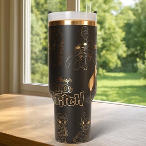 Puede incluir: Vaso negro con ilustraciones doradas de Stitch de Lilo & Stitch de Disney. Tiene una tapa blanca con una banda dorada y una pajita transparente. El texto "Disney's Lilo & Stitch" se muestra en dorado.