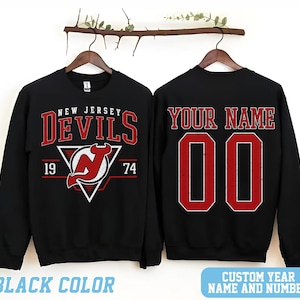 May include: Black sweatshirt with a red New Jersey Devils logo and the text "New Jersey Devils" above the logo. The numbers "19" and "74" are on either side of the logo. The back of the sweatshirt has the text "Your Name" and the number "00" in red.