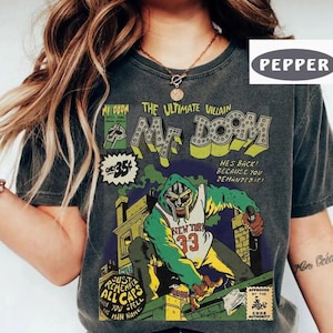 May include: Dark gray t-shirt featuring a comic book-style graphic of MF DOOM. The design includes the text "MF DOOM THE ULTIMATE VILLAIN" and an illustration of a masked figure in a green and white outfit. The shirt also has the text "NEW YORK 33".