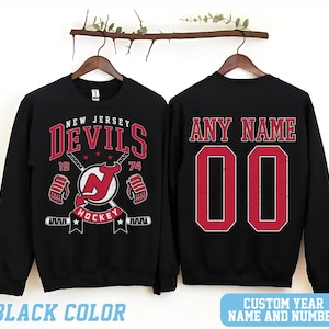 May include: Two black sweatshirts with a New Jersey Devils hockey logo on one and a customizable name and number on the other. The logo features a red and black devil's head with a hockey stick and the words "New Jersey Devils" and "Hockey".