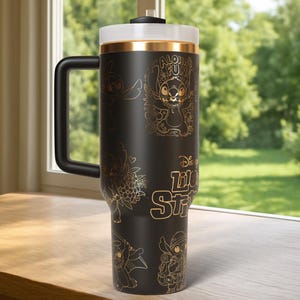 Puede incluir: Taza de viaje negra con ilustraciones doradas de Stitch de Lilo & Stitch. La taza tiene un asa negra, una tapa blanca y una banda dorada. El texto "Aloha Fun" y "Disney Lilo & Stitch" tambi&eacute;n es dorado.