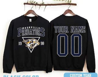 May include: Black sweatshirt with a Nashville Predators logo on the front and a blank space on the back for a custom name and number. The logo features a yellow and blue predator with the team name and years 19 and 98.