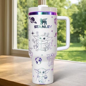 Puede incluir: Vaso Stanley blanco con asa, con ilustraciones de un personaje y gr&aacute;ficos tem&aacute;ticos espaciales. El vaso tiene una tapa blanca y una banda iridiscente p&uacute;rpura y verde. El logotipo de Stanley tambi&eacute;n es visible.
