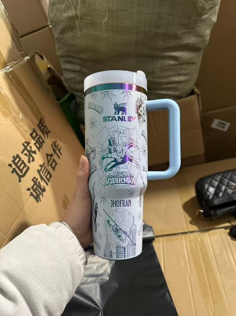 Peut inclure: Un gobelet isotherme blanc Stanley avec des illustrations de Spider-Man et le texte "JHOFRAN". Il poss&egrave;de une poign&eacute;e et un couvercle bleu clair, avec un bord argent&eacute;. Le design comprend des sc&egrave;nes de Spider-Man.
