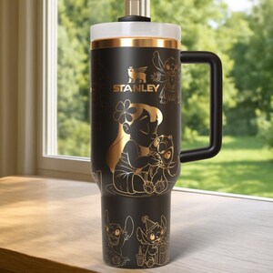 Puede incluir: Vaso Stanley negro con detalles dorados y asa negra. Presenta ilustraciones doradas de personajes de Lilo & Stitch, incluyendo a Lilo y Stitch. El logotipo de Stanley tambi&eacute;n es dorado.