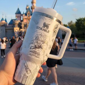 May include: White Stanley tumbler featuring Spider-Man graphics and the Stanley logo. The tumbler has a white handle, lid, and straw. The design includes Spider-Man in action, webs, and cityscapes. Perfect for cold beverages.