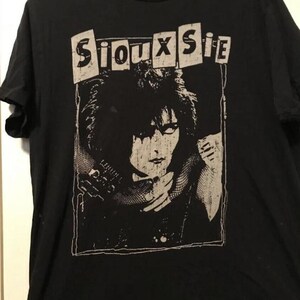 May include: Black t-shirt featuring a black and white graphic of a person with dark hair and the text "Siouxsie" in a rectangular box above the graphic. The t-shirt is made of a soft material.