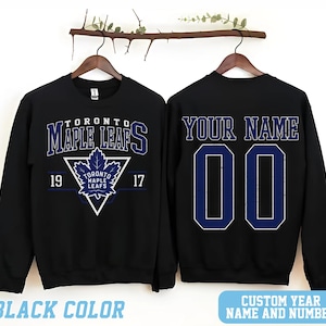 May include: Black sweatshirt featuring the Toronto Maple Leafs logo in white and blue. The front displays the team name and a maple leaf emblem. The back has space for a custom name and number in blue.