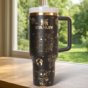 Puede incluir: Vaso Stanley negro con tapa blanca y asa negra. El vaso presenta ilustraciones doradas de un personaje y dise&ntilde;os tem&aacute;ticos espaciales. El logotipo de Stanley se muestra en dorado. El vaso est&aacute; sobre una superficie de madera.