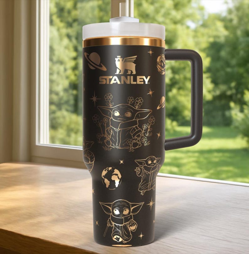 Puede incluir: Vaso Stanley negro con tapa blanca y asa negra. El vaso presenta ilustraciones doradas de un personaje y dise&ntilde;os tem&aacute;ticos espaciales. El logotipo de Stanley se muestra en dorado. El vaso est&aacute; sobre una superficie de madera.