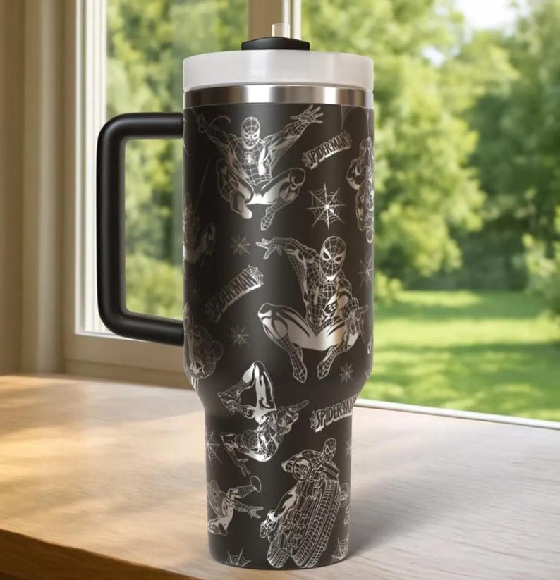 May include: Black travel mug featuring a Spider-Man design. The stainless steel tumbler has a black handle, a white lid, and a silver rim. The design includes Spider-Man in various action poses and the word "Spider-Man".