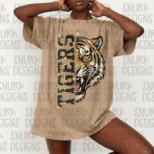 May include: A tan-colored t-shirt featuring a detailed graphic of a tiger's face with the word "TIGERS" in vertical, sparkling letters. The tiger design incorporates gold and black accents.