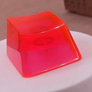 May include: Five bright pink keycaps, made of translucent plastic, are arranged on a white surface. One keycap is on a small white pedestal.